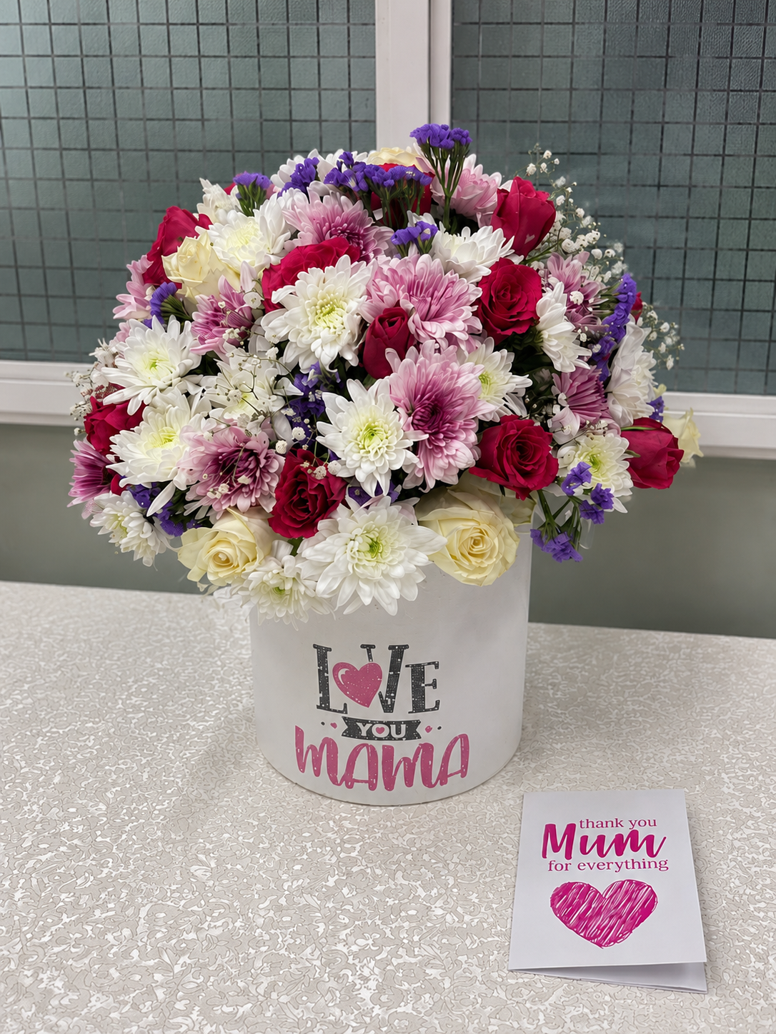 A bouquet of flowers in a box with 'Love You Mama' text, placed on a table. By Simona Flowers and Gifts