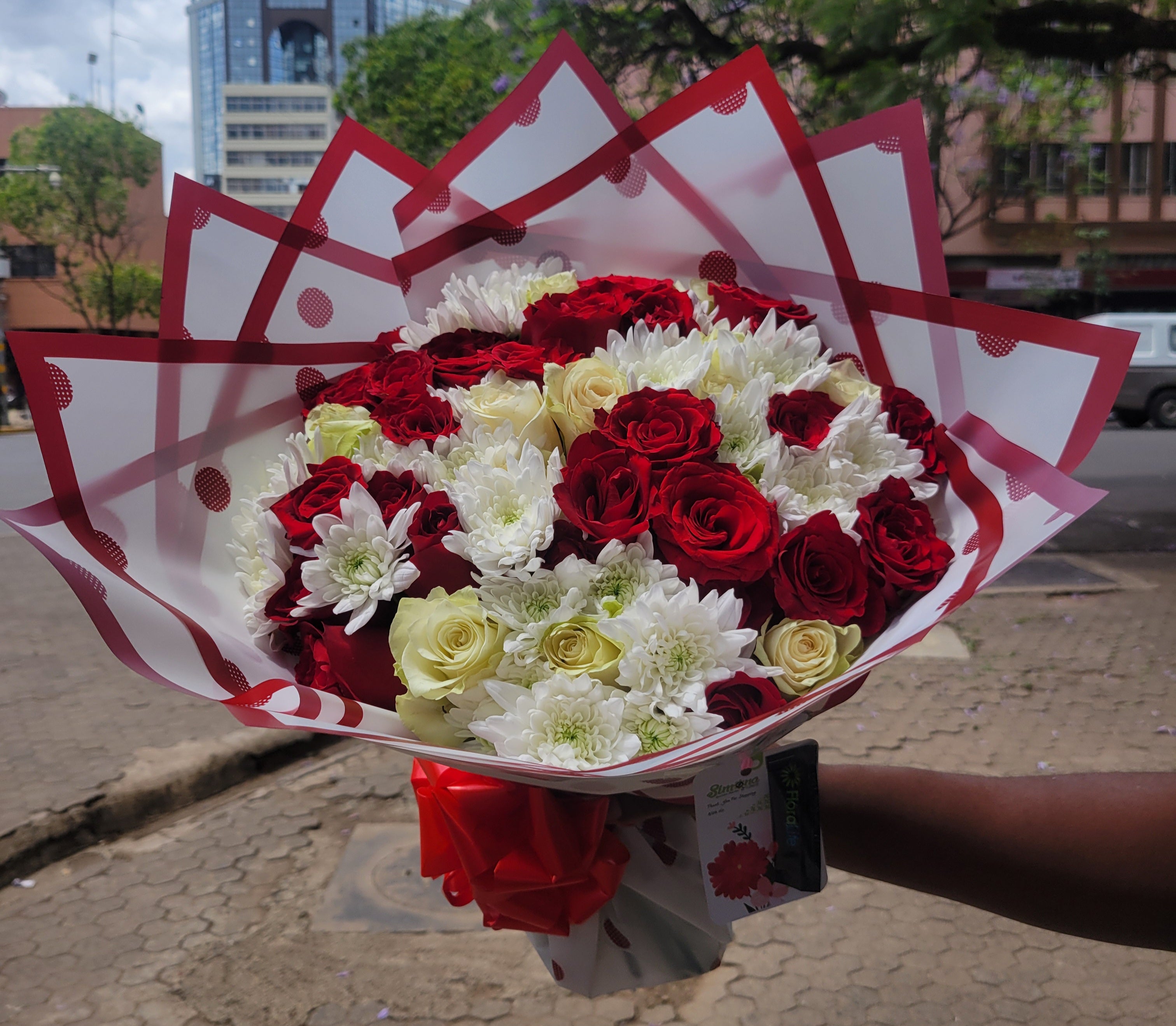 Bouquet of red, white, and yellow flowers in a decorative paper wrap held by a person outdoors.
