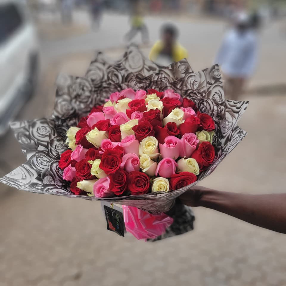 A bouquet of pink, red, and white roses held by a person on a blurred street background. By Simona Flowers Kenya