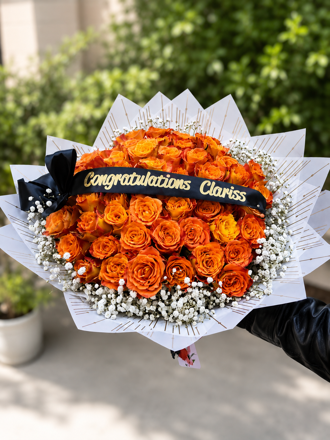Bouquet of orange roses with a black bow and 'Congratulations Clariss' message, set against a blurred green outdoor background. By Simona Flowers.